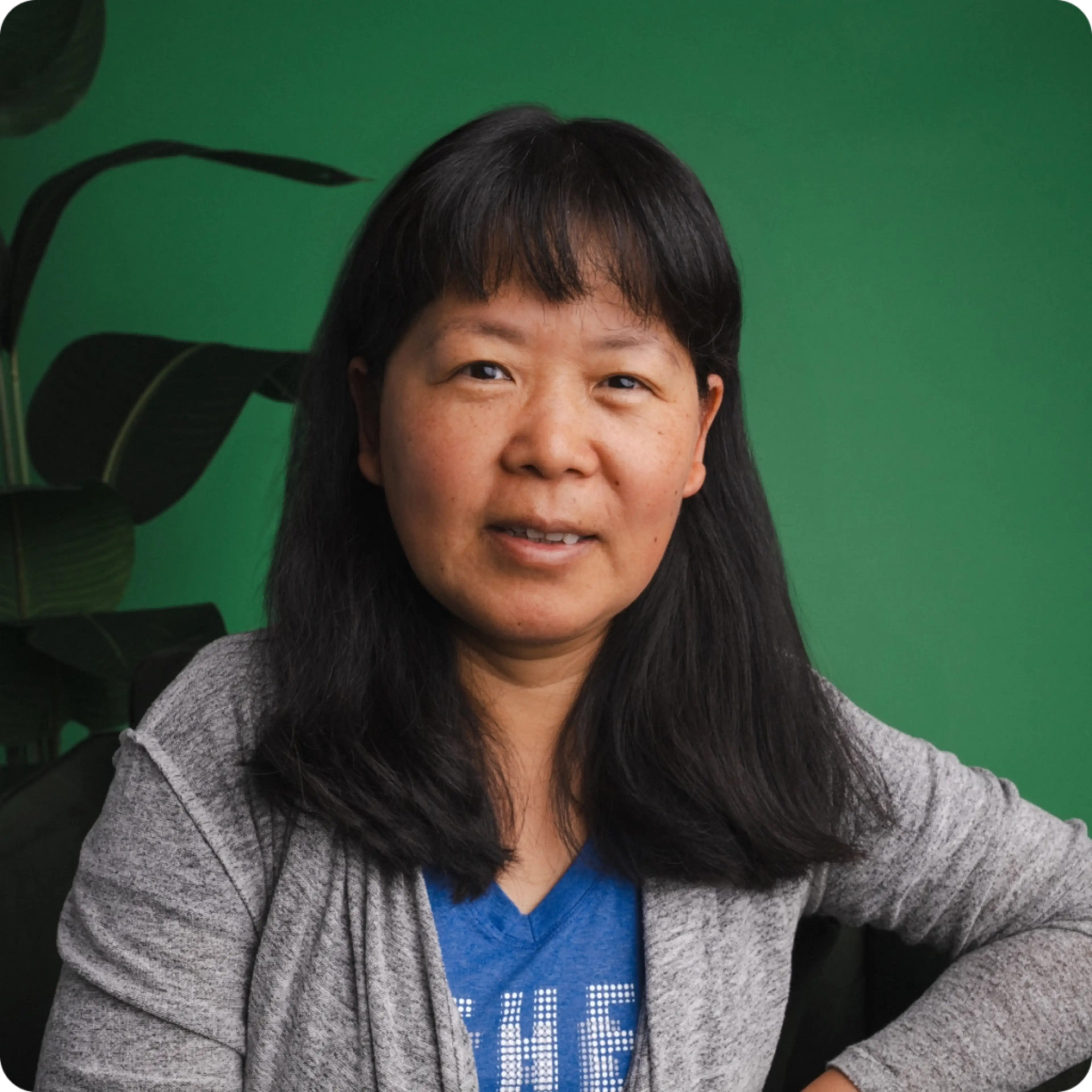 A woman with long black hair smiles softly, wearing a gray cardigan and blue shirt, posed before a green wall with a plant.