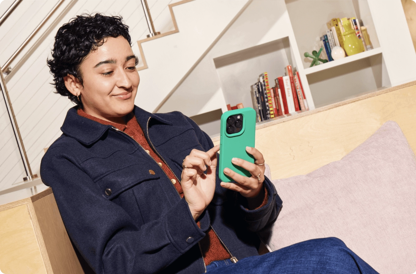 Person with curly hair in navy jacket using the Chime mobile app on a cell phone, sitting in modern room with bookshelves.