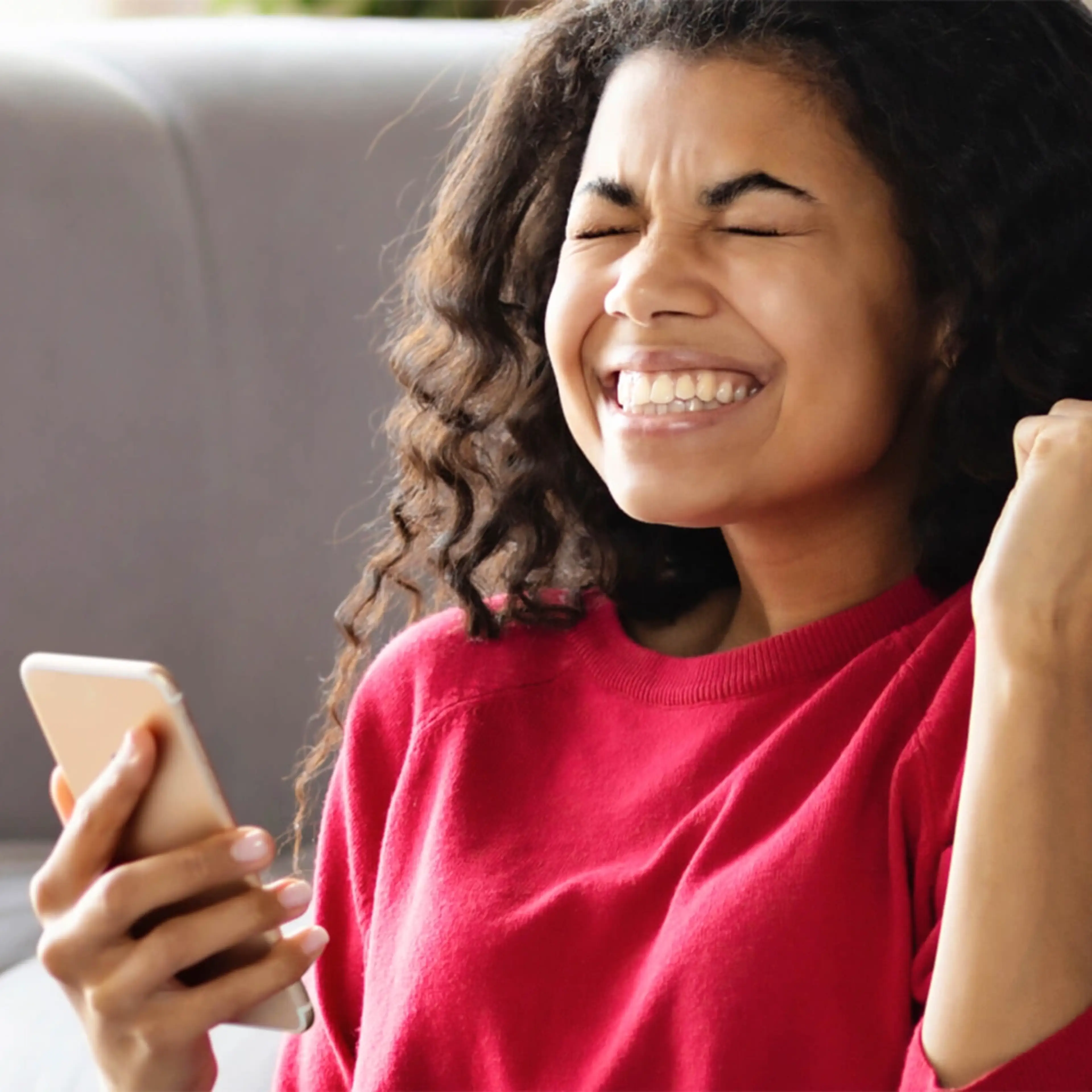 Person with curly hair in red sweater laughing joyfully while looking at smartphone with raised fist in celebration.