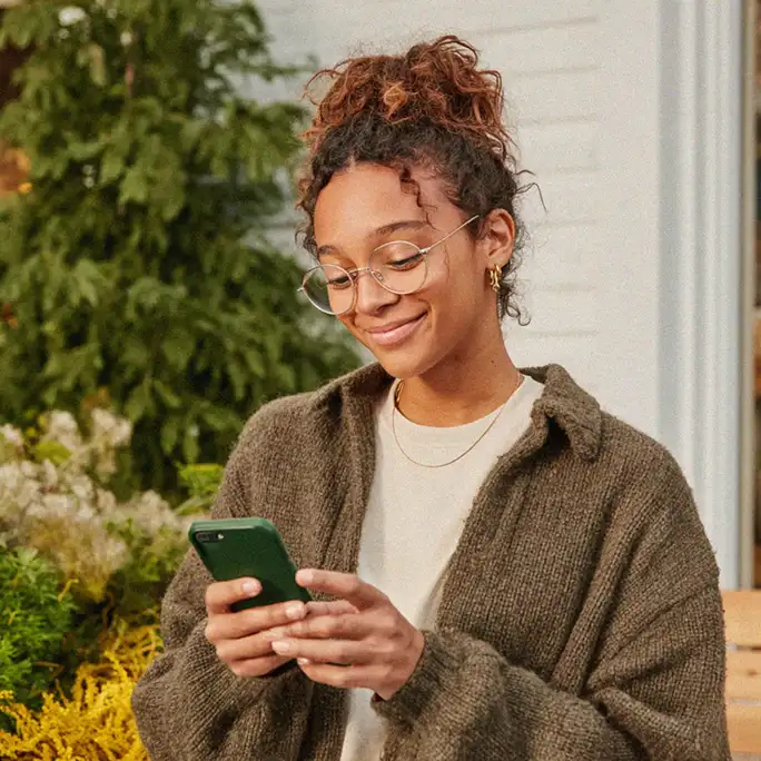 A smiling young woman with curly hair and glasses looks at her green smartphone outdoors near lush greenery.