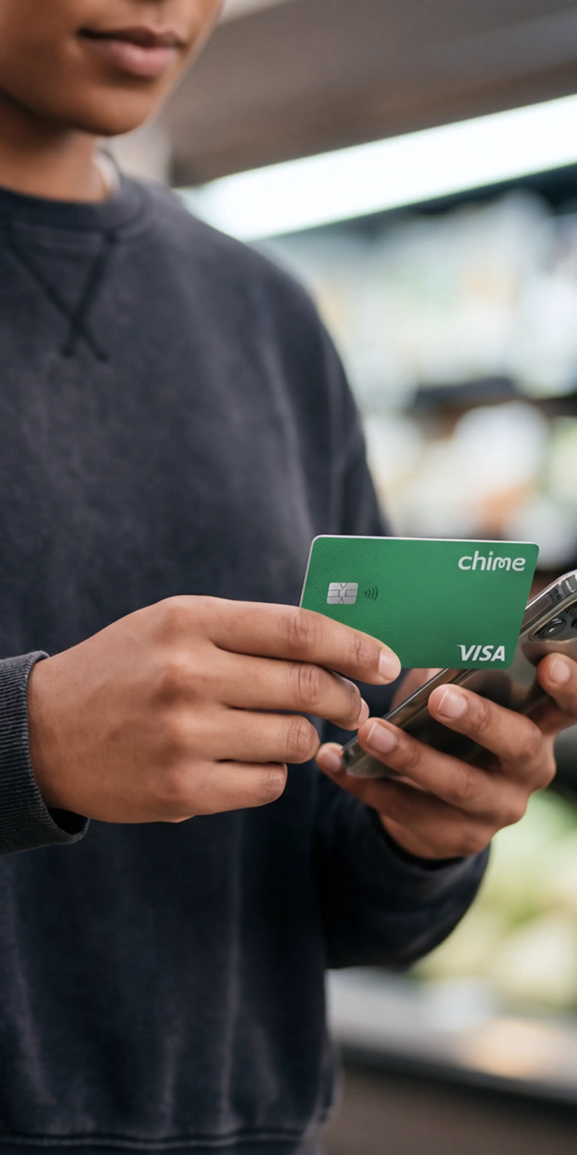 Person in a gray sweatshirt holding a green Chime Visa card and a smartphone at a payment terminal in a store.