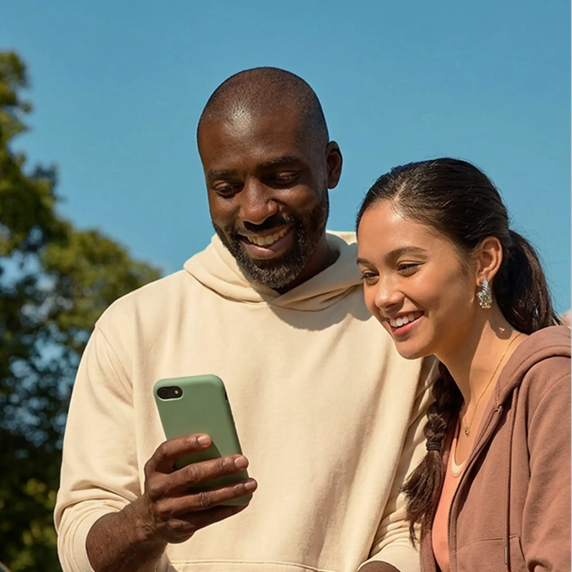 Two smiling adults outdoors looking at a green smartphone together under a clear blue sky with trees in the background.