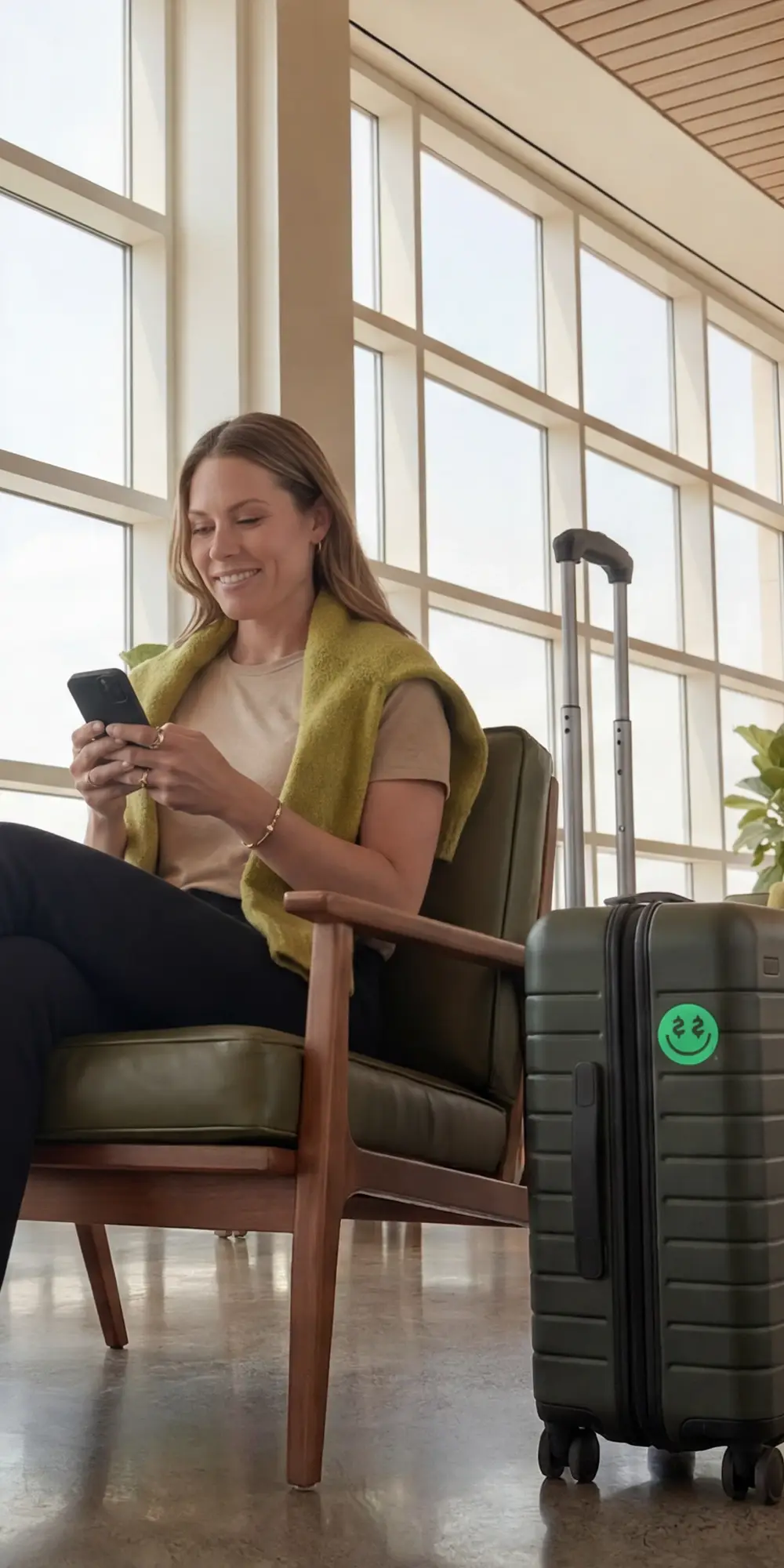 Woman smiling at her phone in an airport lounge, seated beside a dark carry-on suitcase with a green smiley face sticker.