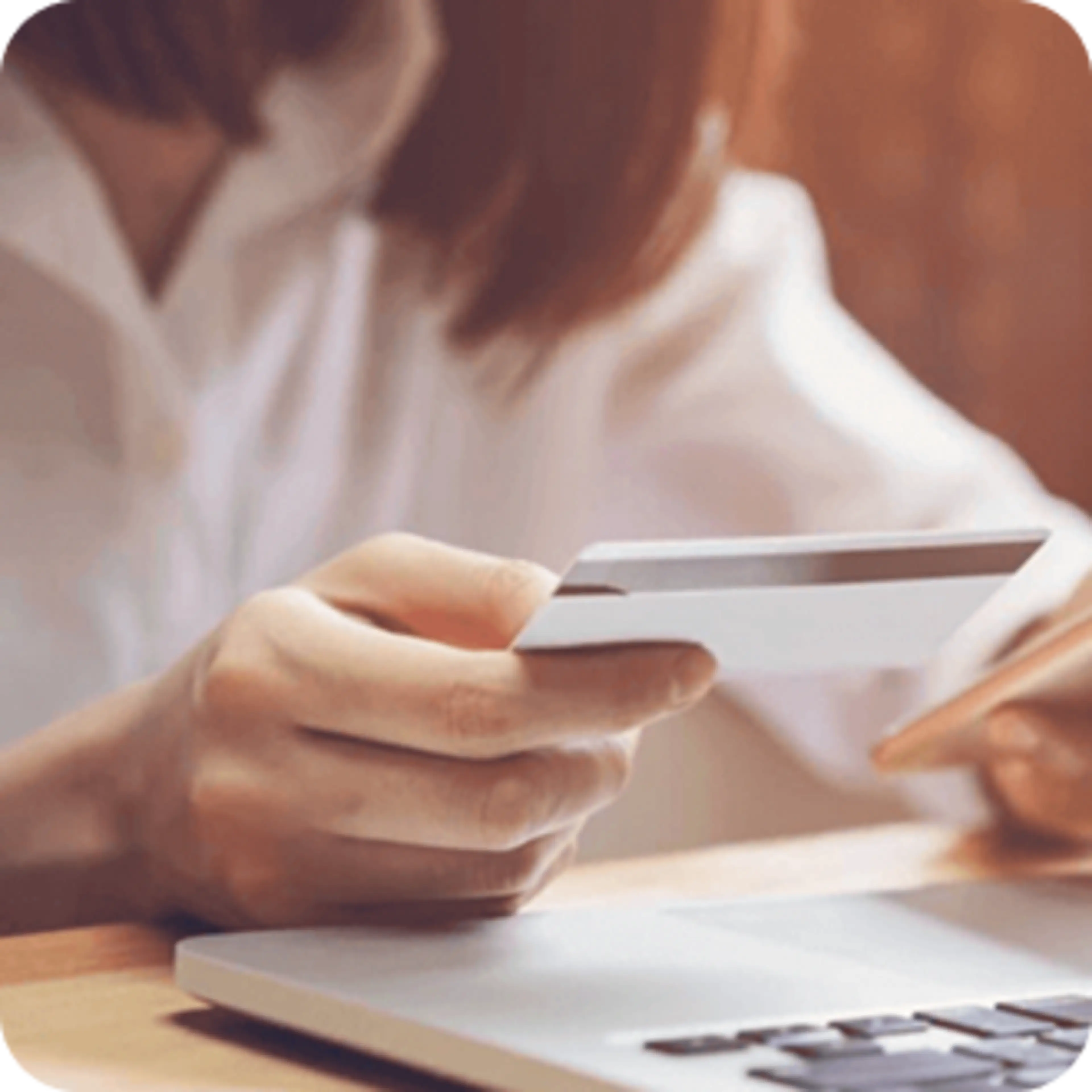 Person in white shirt holding a credit card while seated at a desk with a laptop visible.