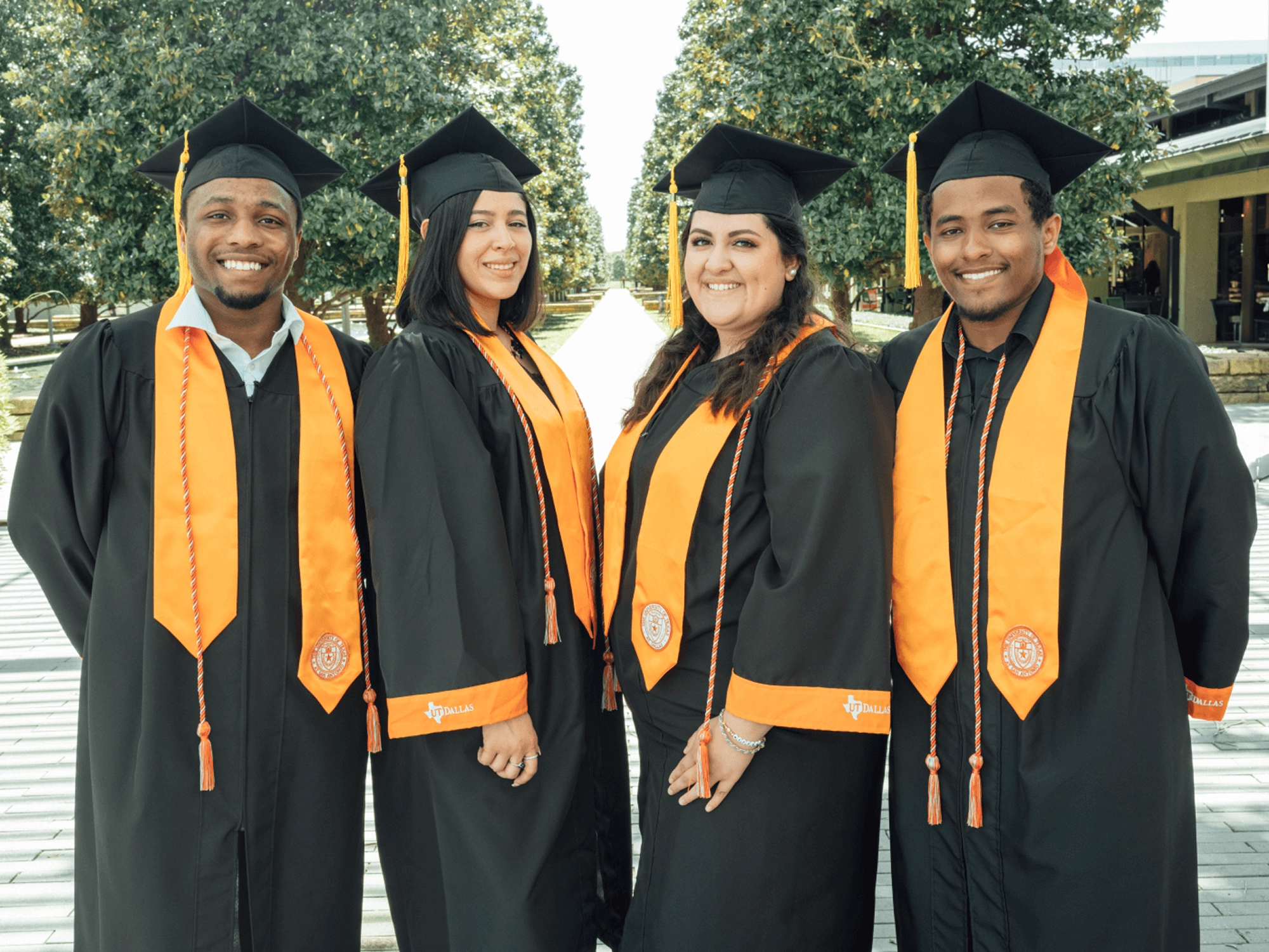 Four graduates in black caps and gowns with orange stoles on a campus walkway.