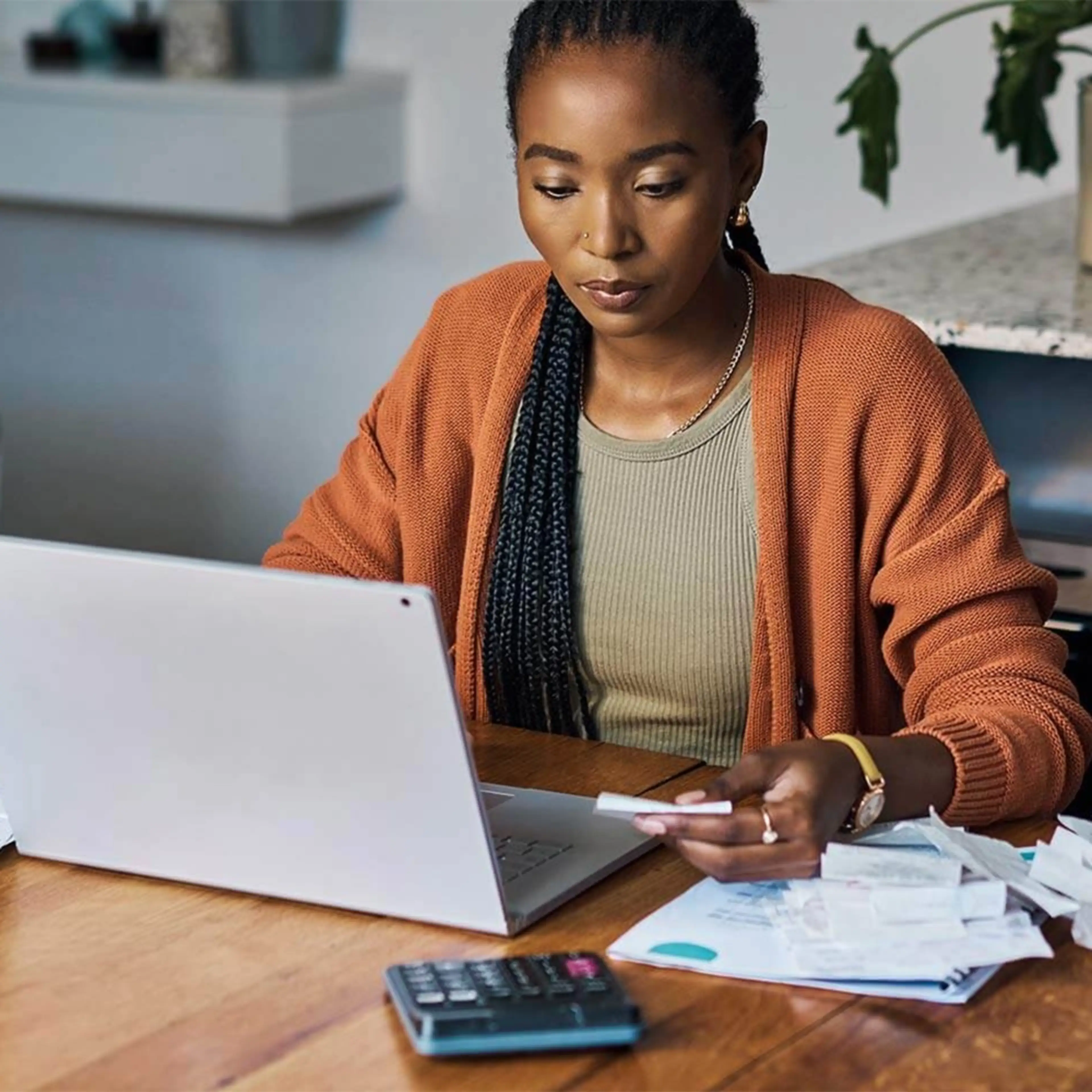 Woman working on laptop with papers and calculator on wooden desk in home office setting.