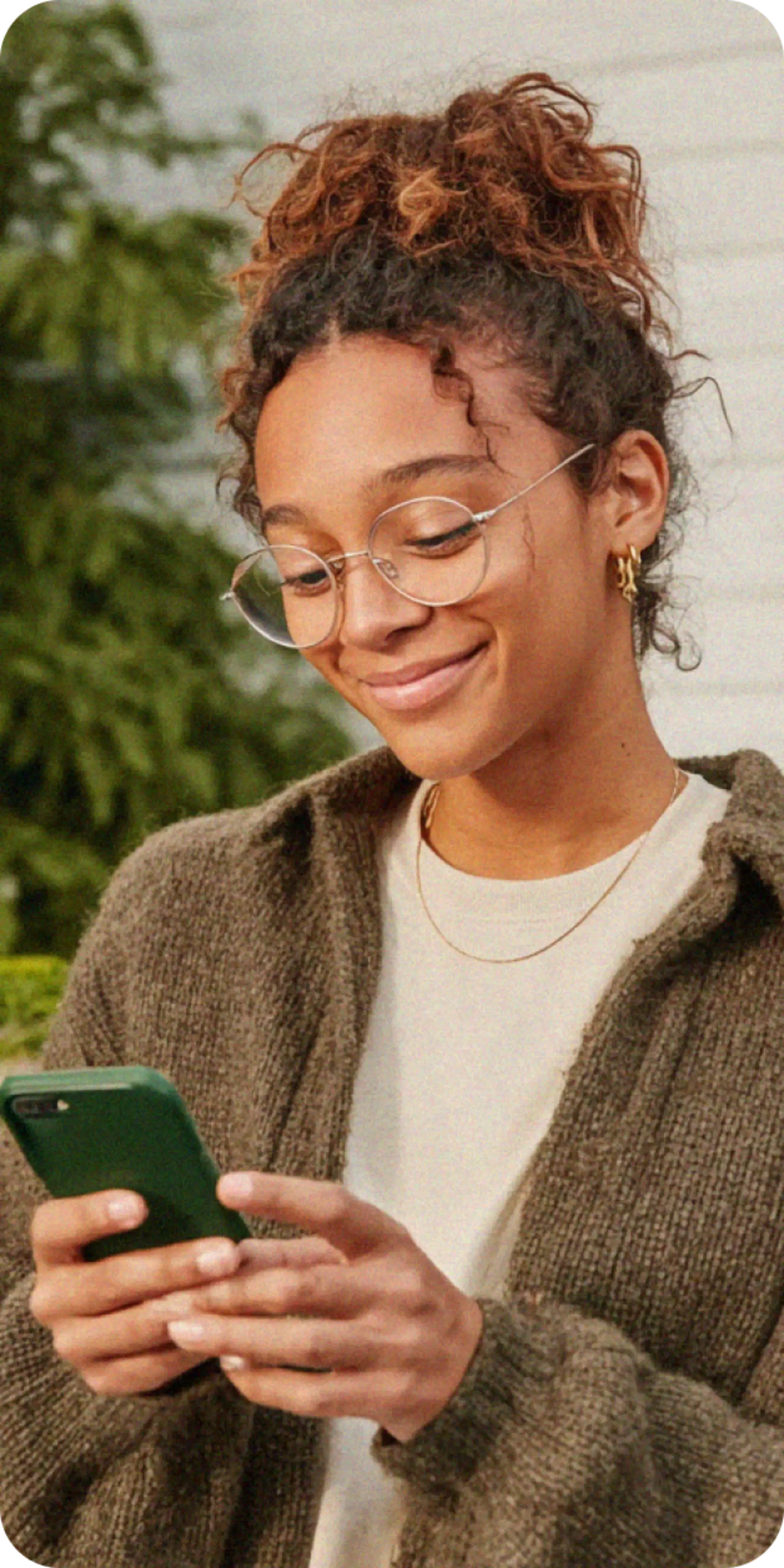 A young woman with curly hair and round glasses smiles while looking at her green smartphone outdoors.
