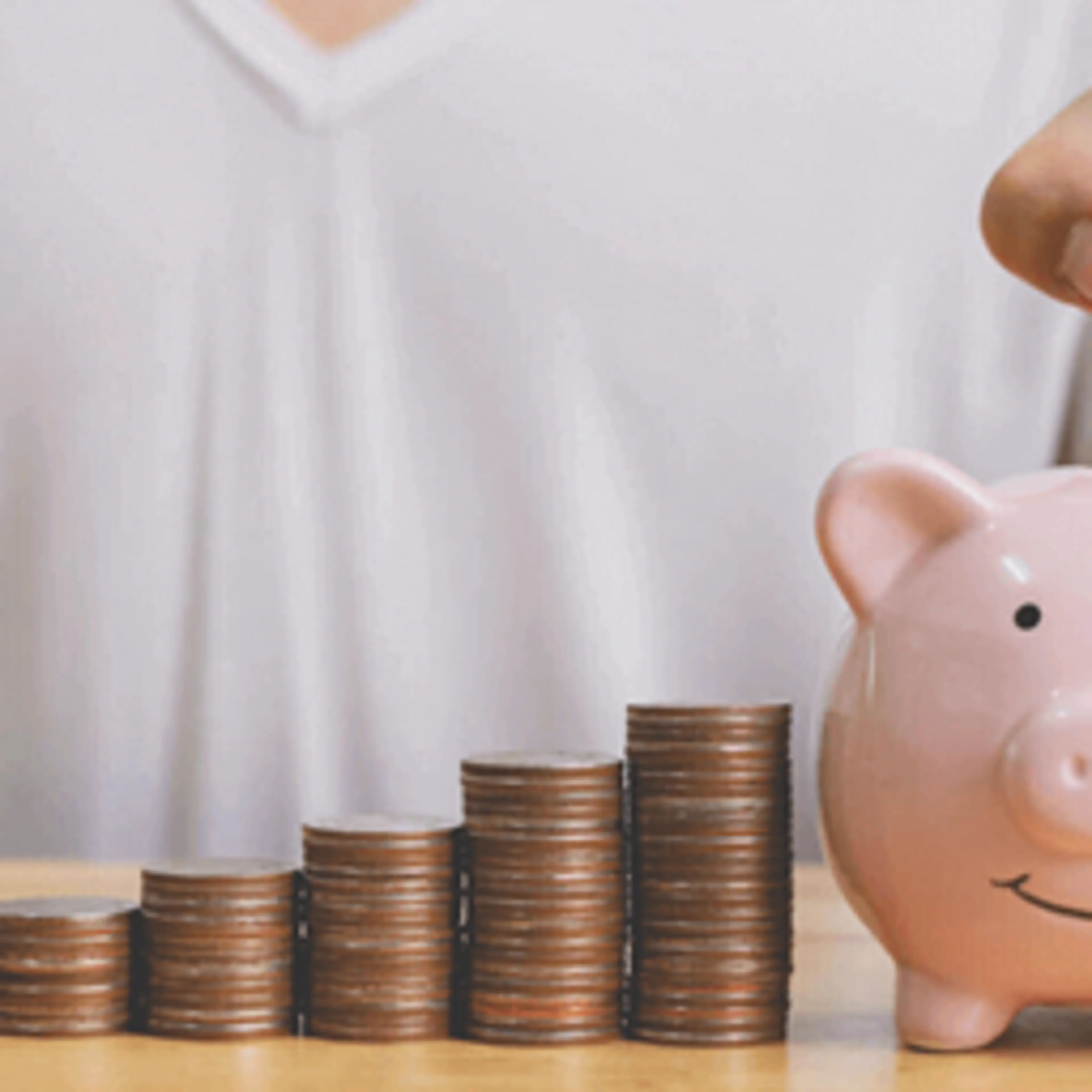 Person in white shirt placing coin into pink piggy bank beside ascending stacks of coins showing savings growth.