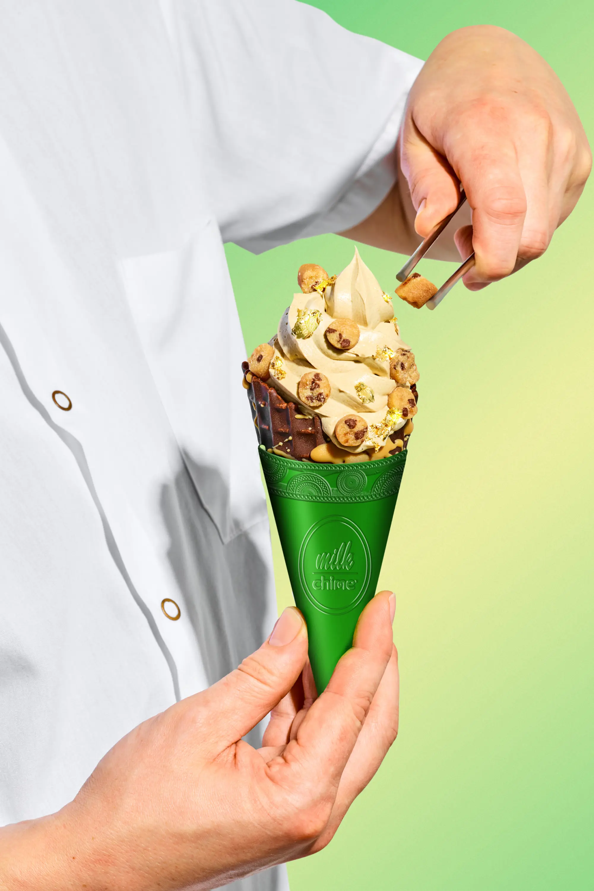 A person in a white coat holds a green Milk Chime cone topped with swirled soft serve, cookie dough balls, and gold leaf.