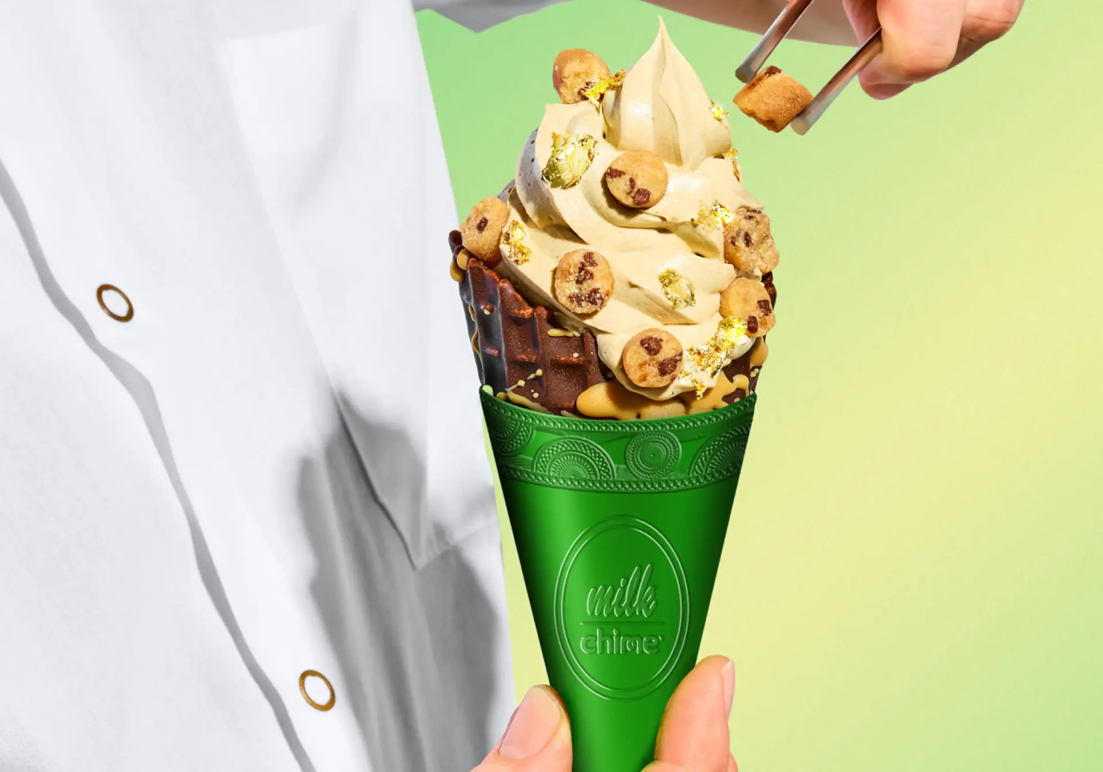 A person in a white coat holds a green Milk Chime cone topped with swirled soft serve, cookie dough balls, and gold leaf.