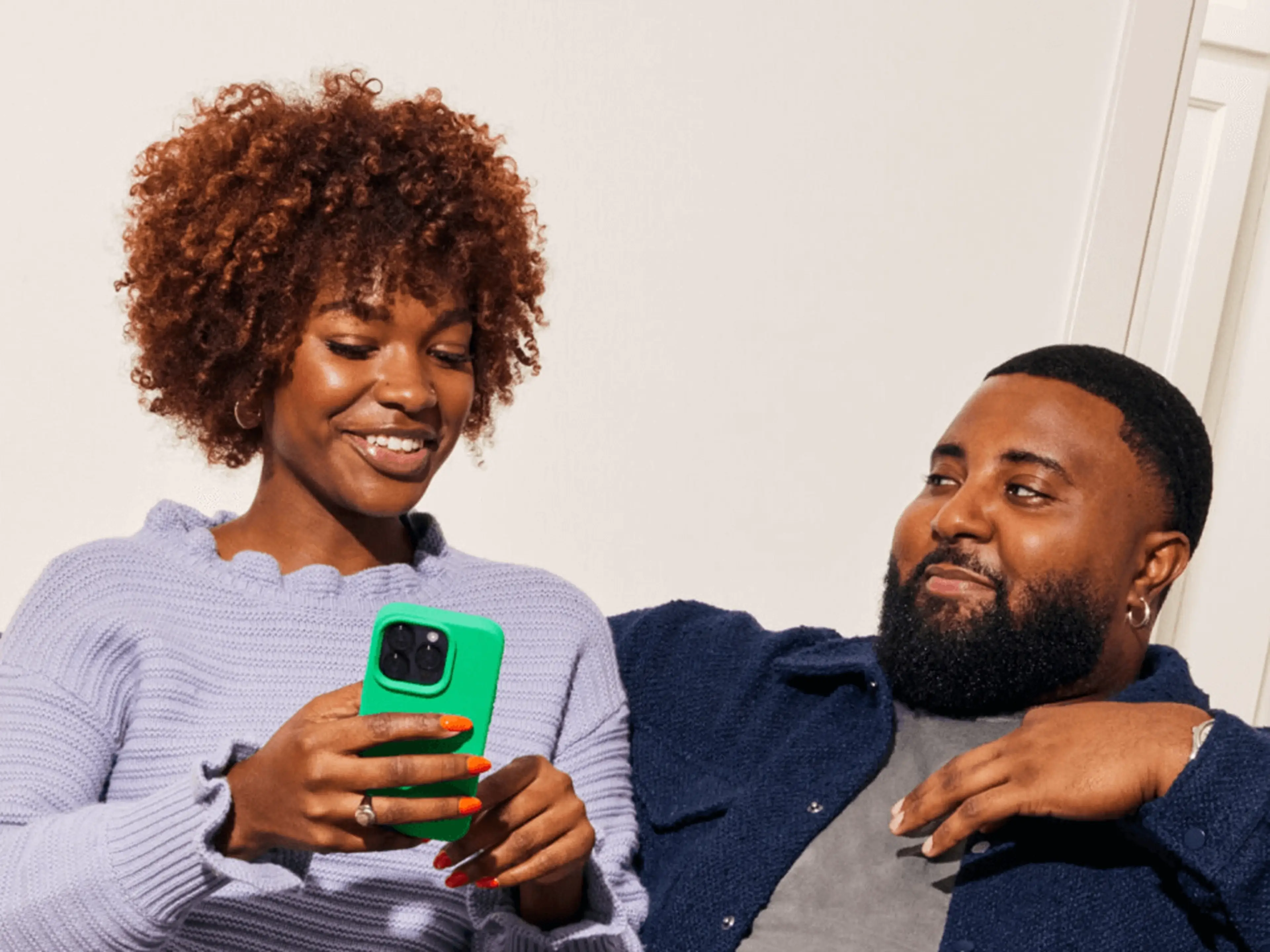 Young woman in lavender holds a phone in green case while man looks on.
