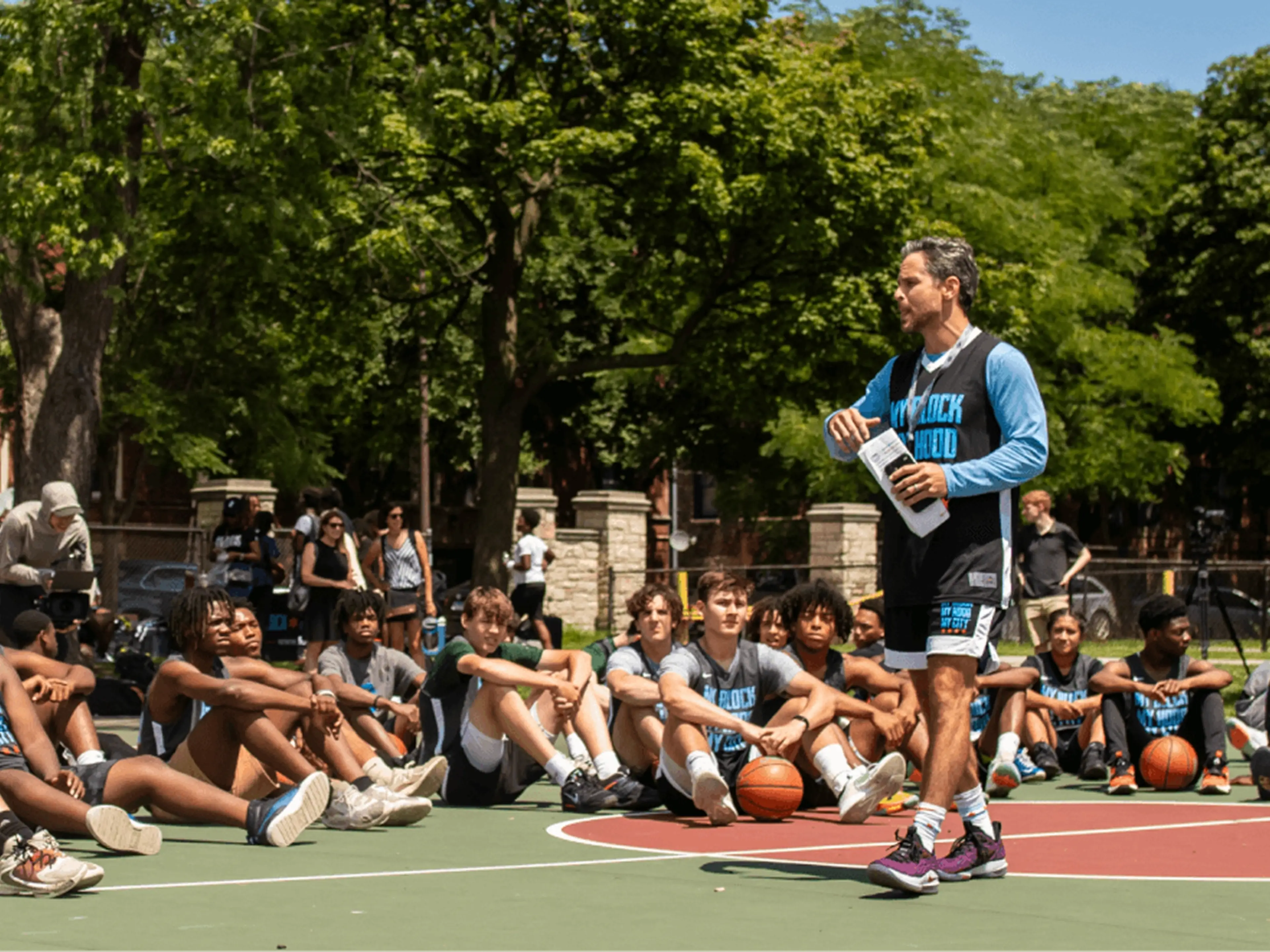 Man in basketball jersey speaks to teenagers on an outdoor community basketball court.