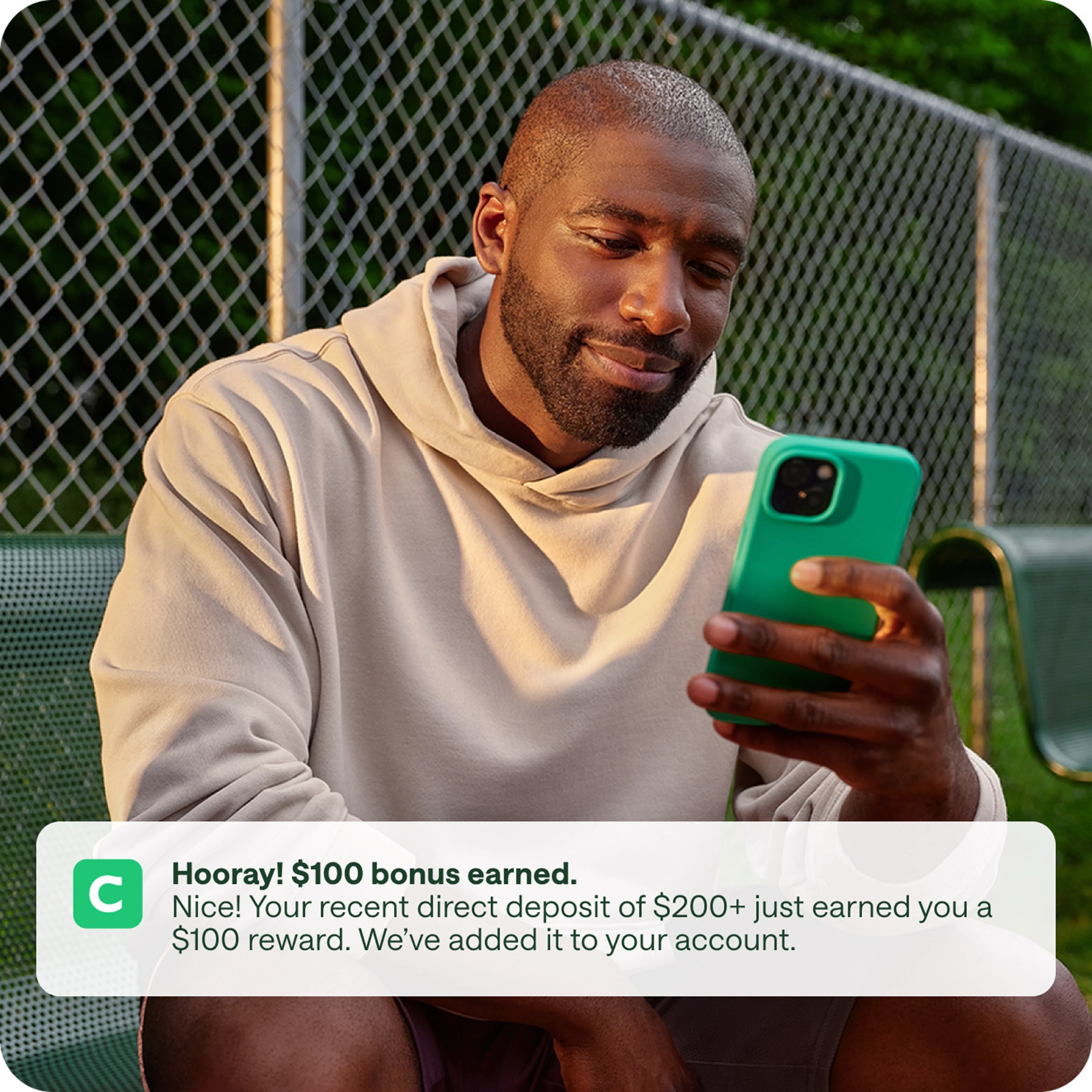 Man sitting on bench receiving $100 Chime bonus on phone after direct deposit, with Chime banking app notification on screen. The screen says, “Hooray! $100 bonus earned. Your recent direct deposit of $200+ just earned you a $100 reward. We've added it to your Checking Account.”
