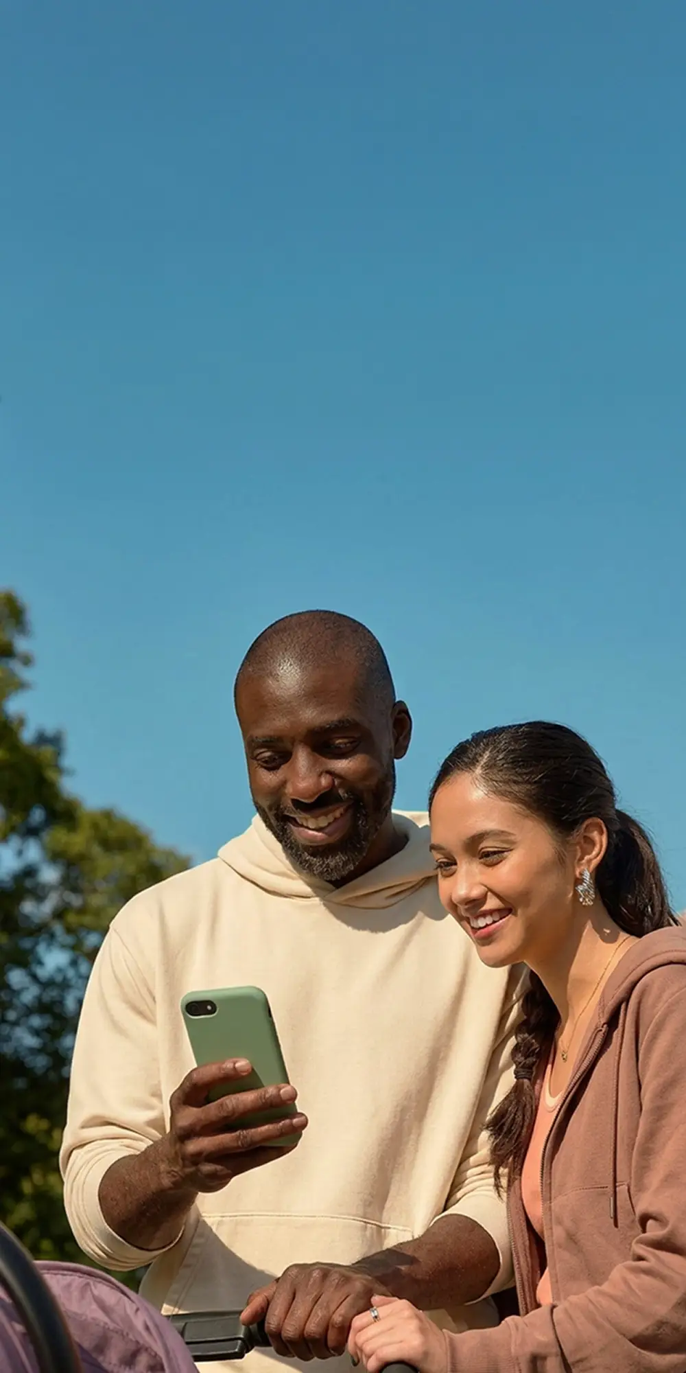 Two smiling adults outdoors looking at a green smartphone together under a clear blue sky with trees in the background.