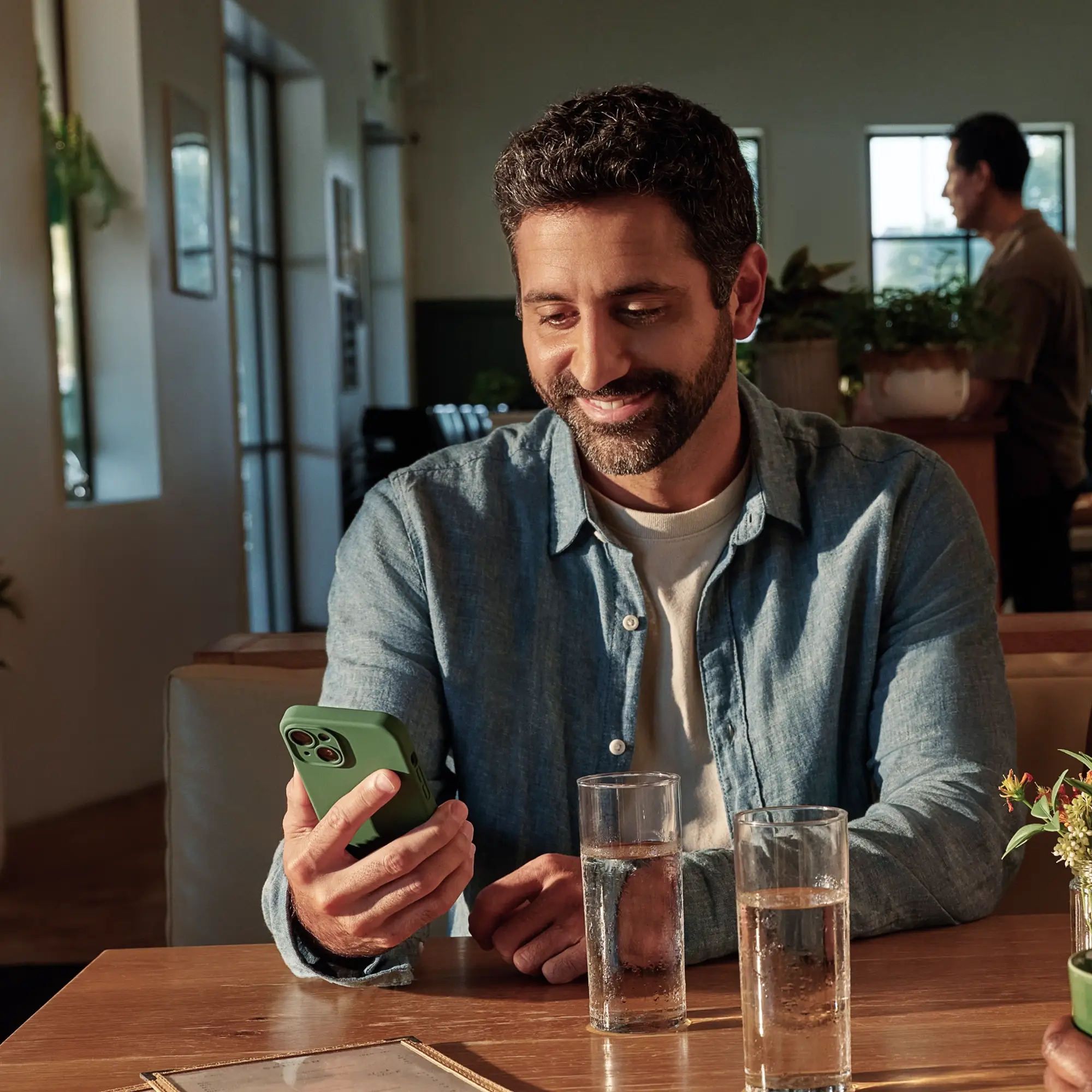A smiling bearded man in a blue shirt looks at his green-cased smartphone at a table with two water glasses.