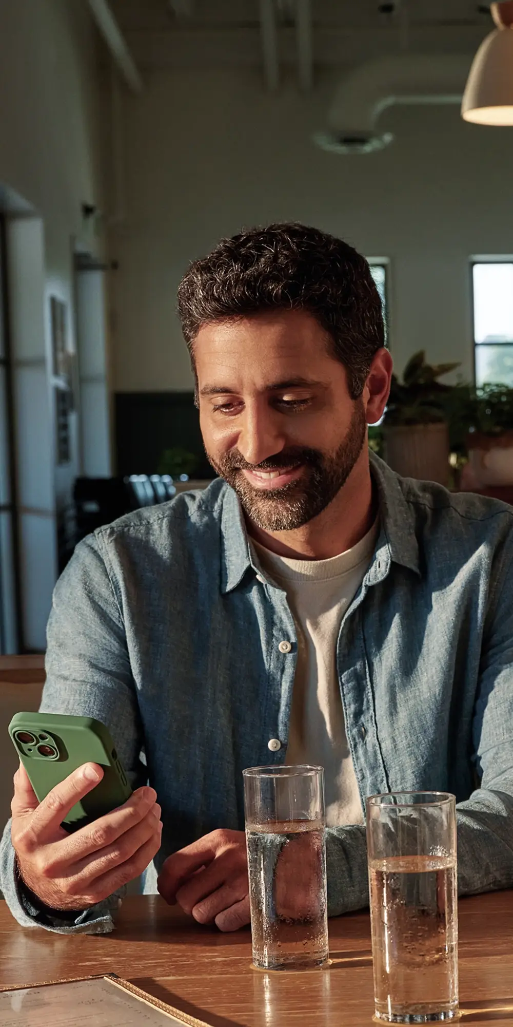 A smiling bearded man in a blue shirt looks at his green-cased smartphone at a table with two water glasses.