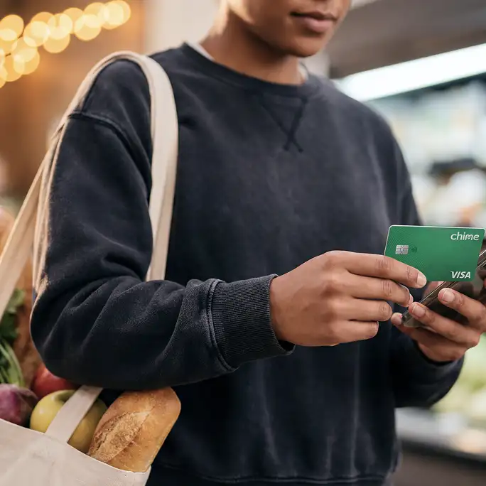 Person holding a green Chime Visa card and smartphone at a grocery store, carrying a tote bag filled with fresh produce and bread.