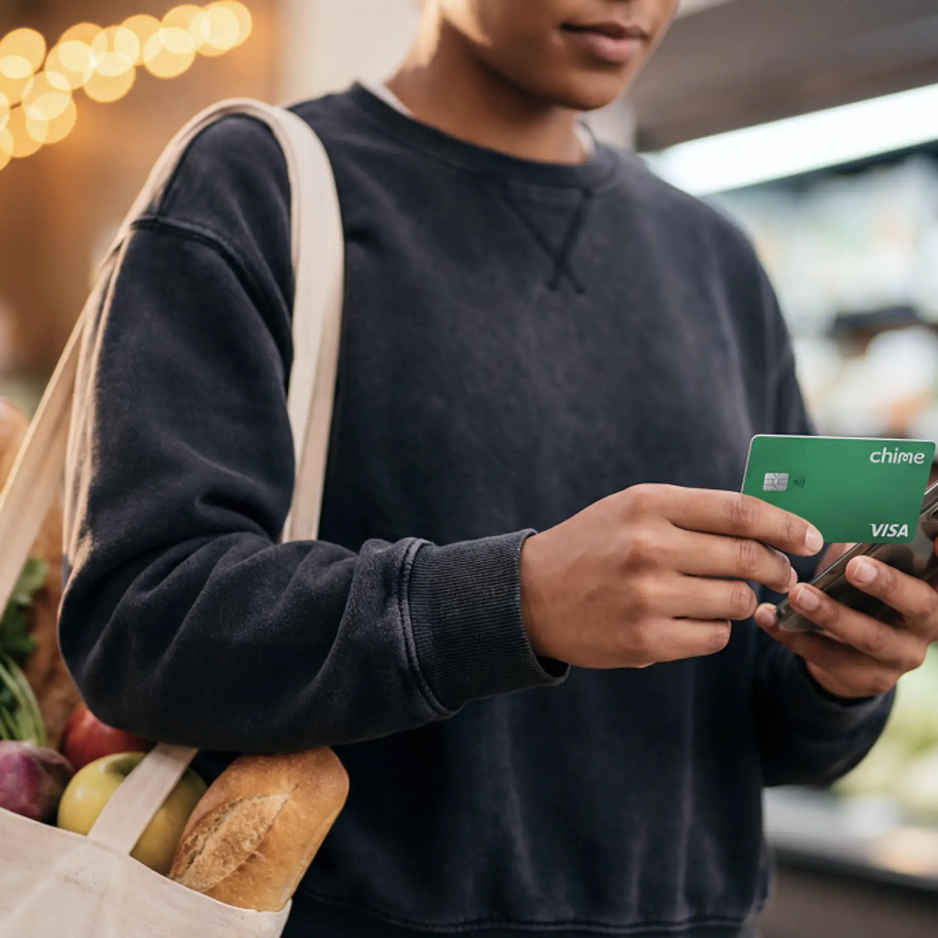 Person holding a green Chime Visa card and smartphone at a grocery store, carrying a tote bag filled with fresh produce and bread.