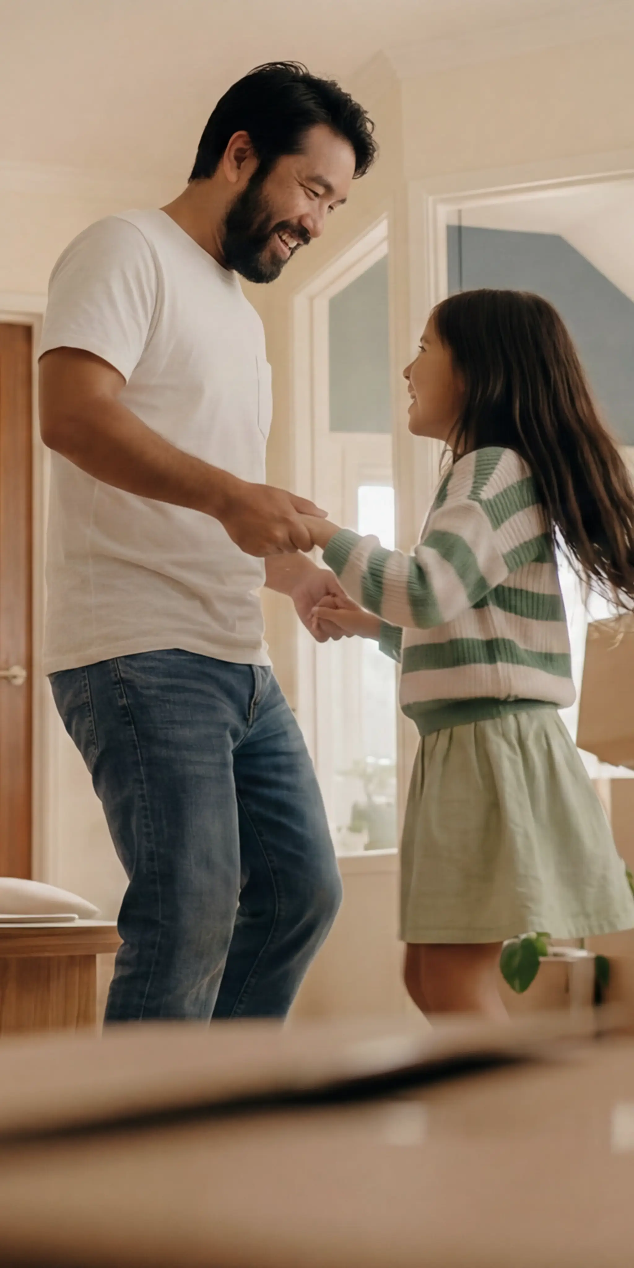 A smiling father in a white t-shirt and jeans dances with his young daughter in a bright, cozy home.