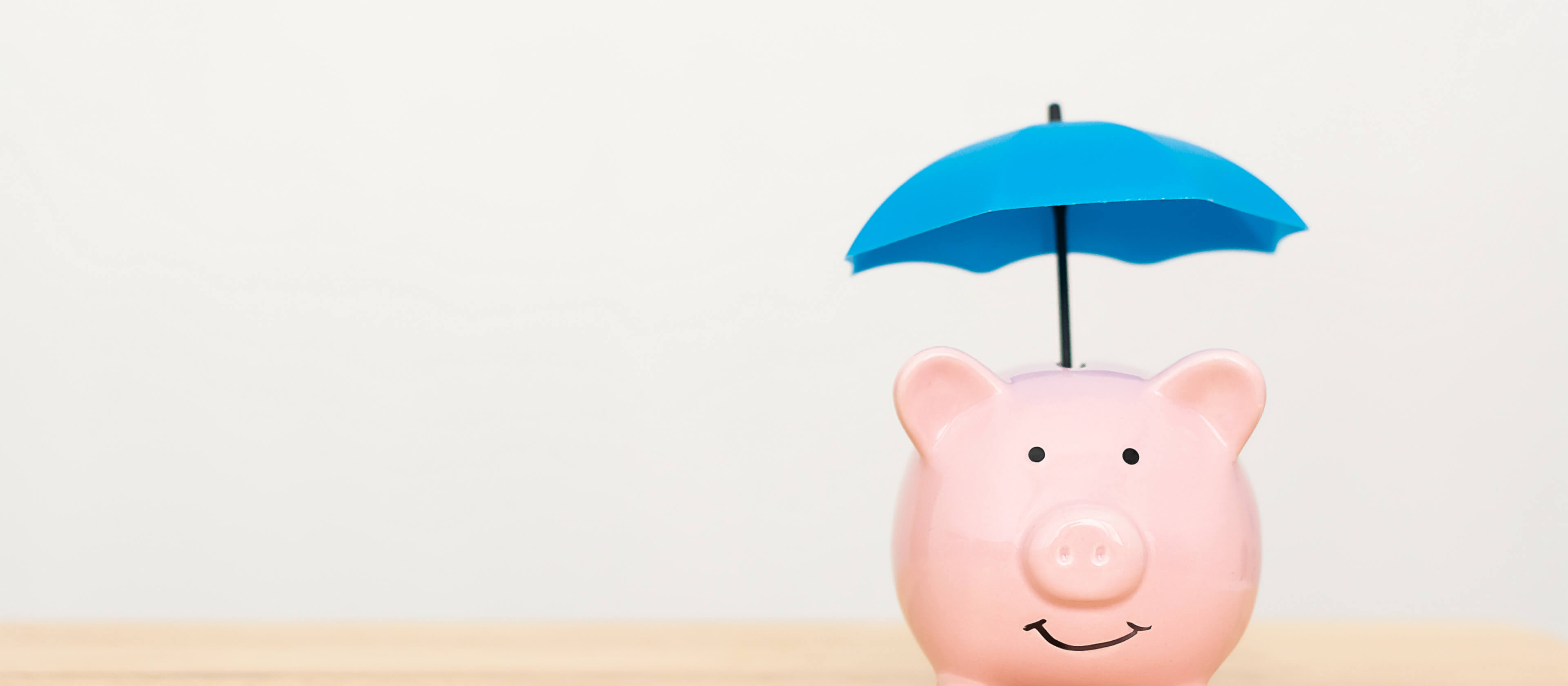 A piggy bank with a blue umbrella above it sits on a wooden table.