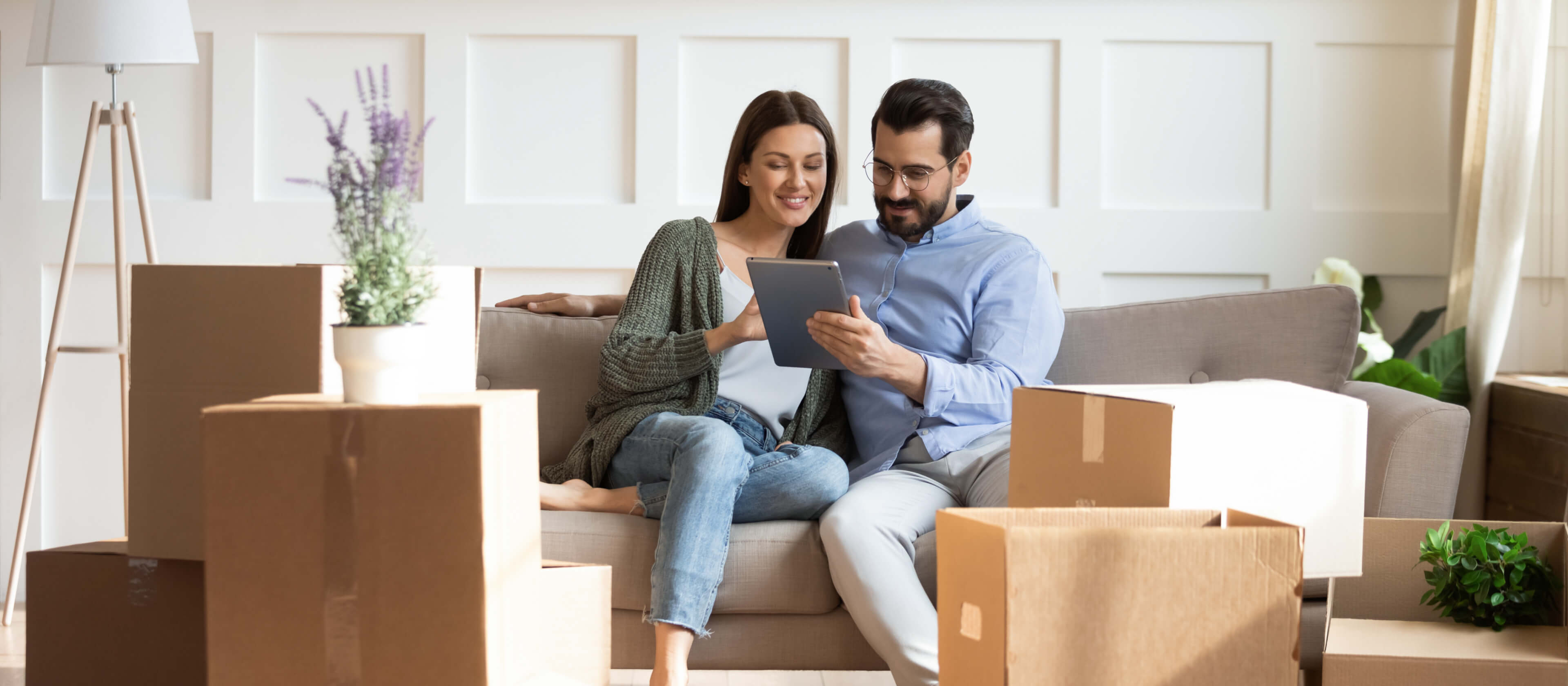 A young couple sits on their couch with boxes around them and reviews an iPad.