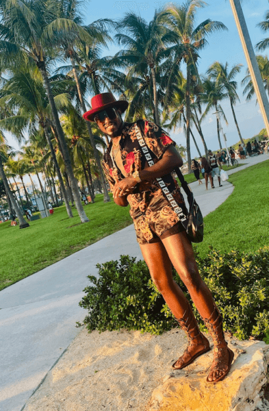 A man stands on a rock next to the sidewalk. He is wearing a two piece black and red outfit with a red hat, smiling.