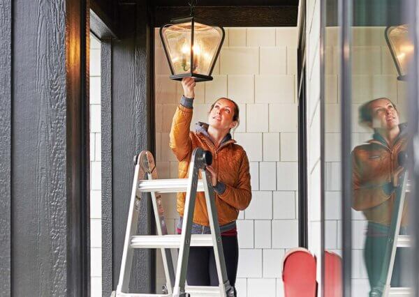 A woman stands on a ladder to replace a light bulb above.