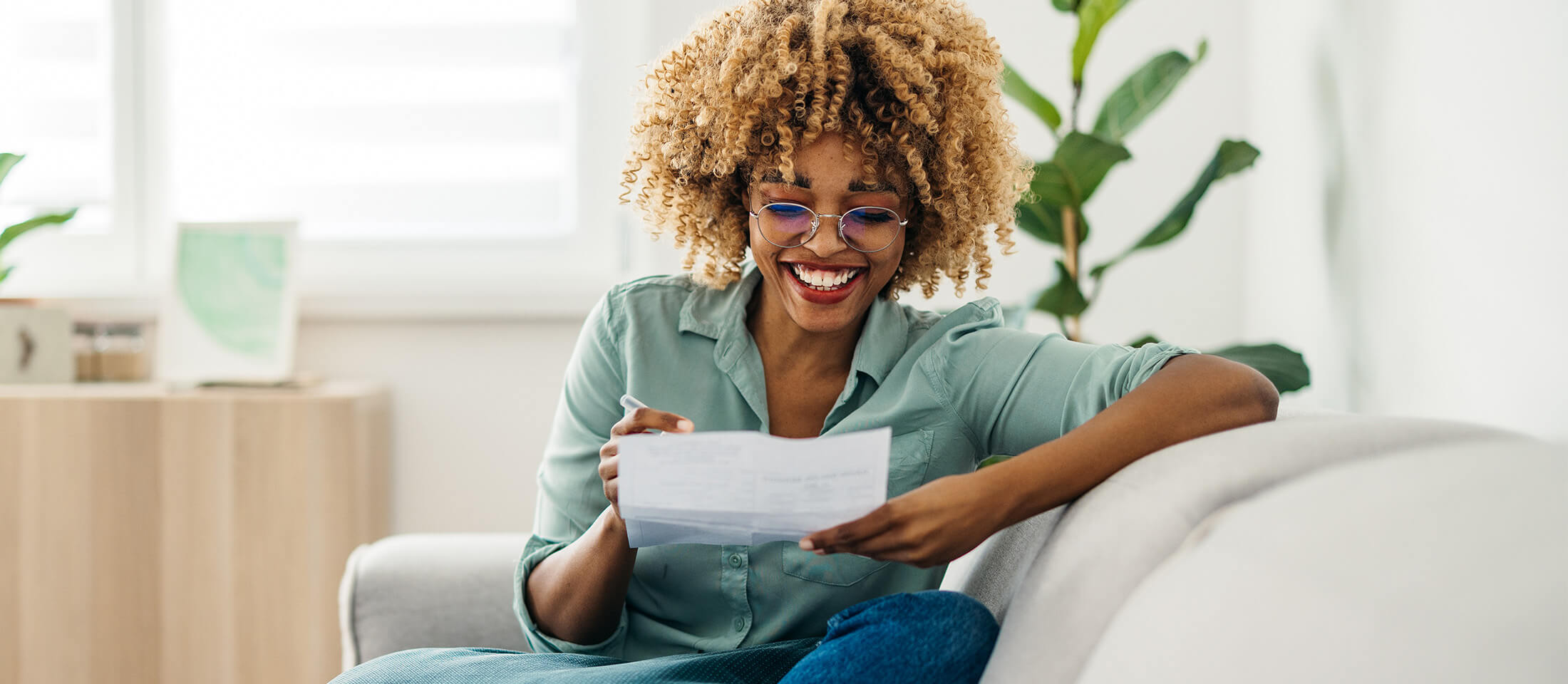 A woman sits on her couch smiling while reading a letter.