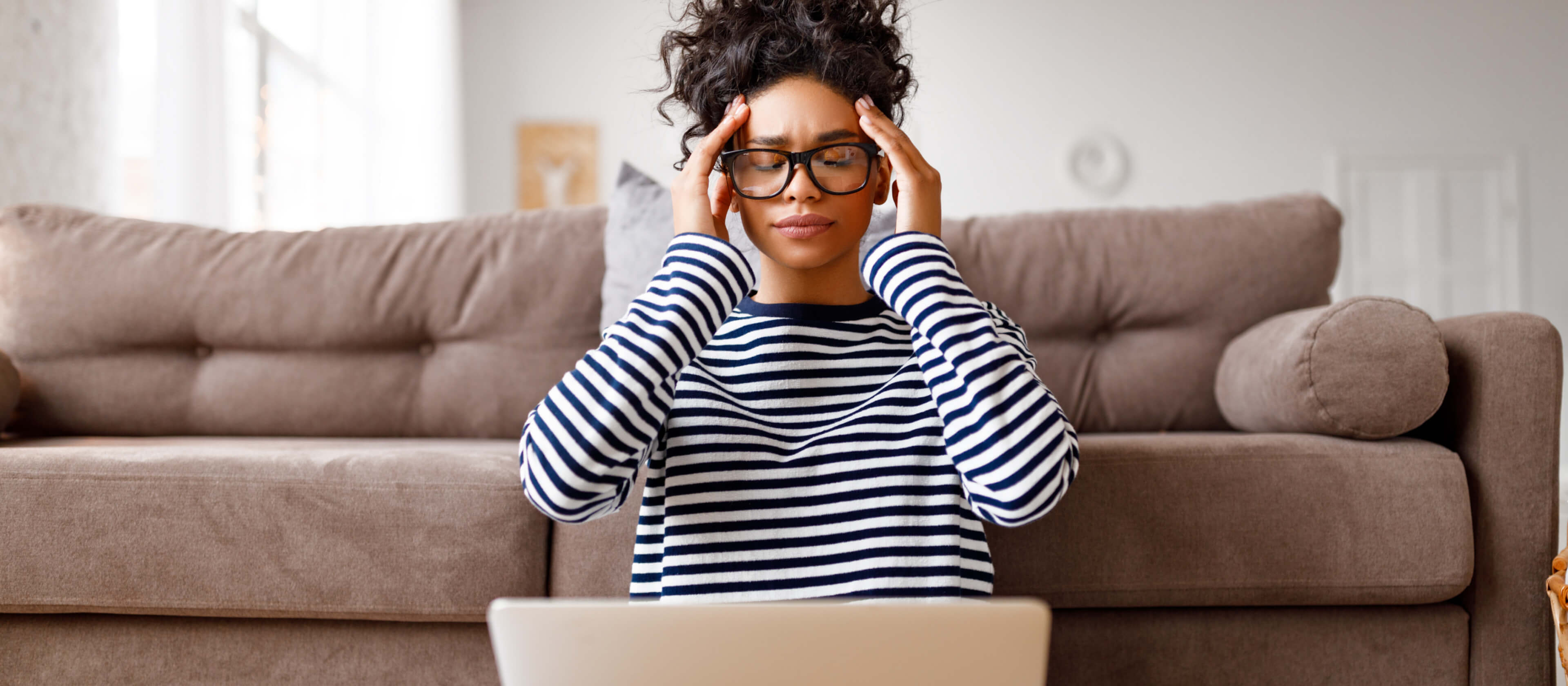 A young woman sits against her couch with her laptop in her lap. She presses her hands to her temples, visibly stressed.